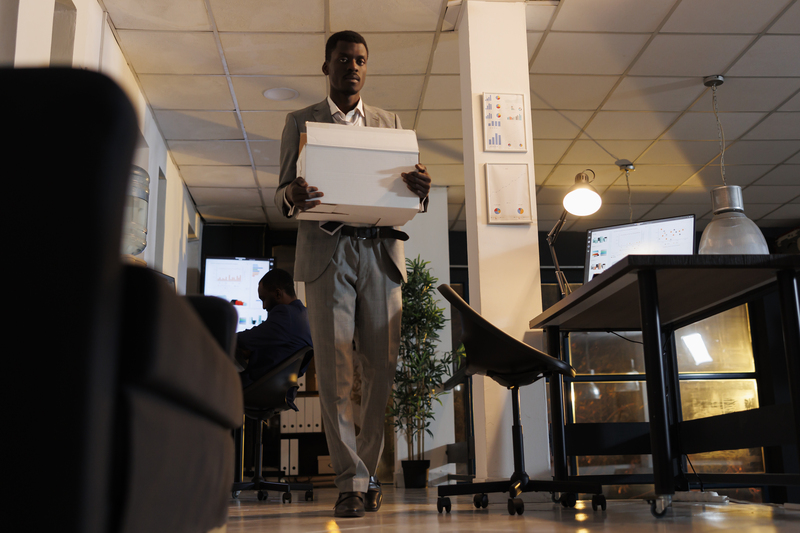 African american entrepreneur holding cardboard box with his belongings after being fired from business startup office. Advisor agent quitting corporate job late at night. Dismissal concept