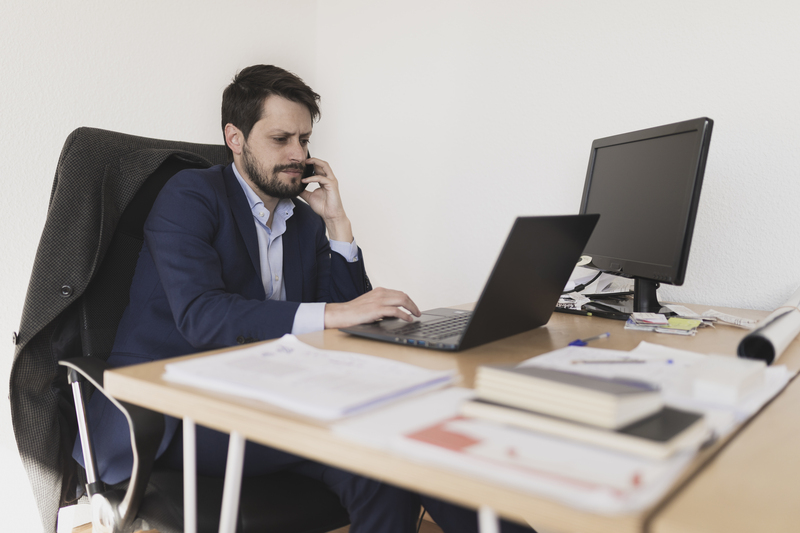 Concentrated young male talking on mobile phone and browsing on laptop at table in office