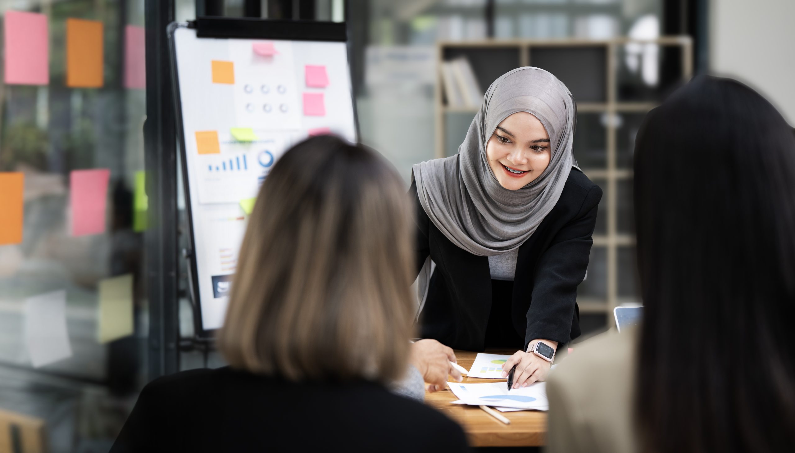 Confident Asian muslim female team leader wearing hijab discussing project statistics with colleagues, standing at table in boardroom
