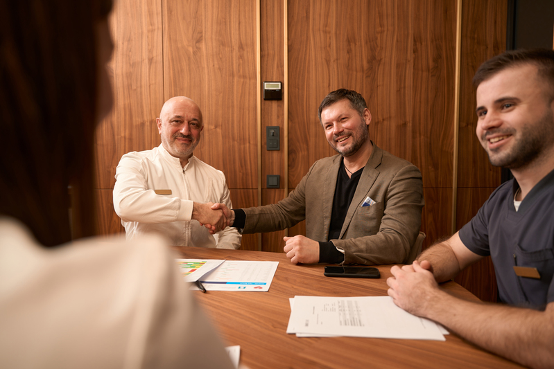 Adult physician sitting at table with colleagues and shaking hands with patient in the office