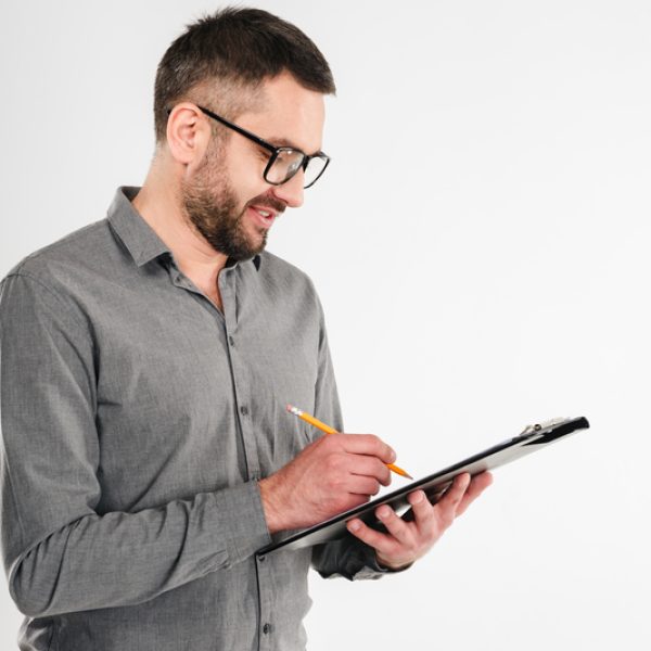 Image of handsome businessman standing isolated over white background. Looking aside holding clipboard writing notes.