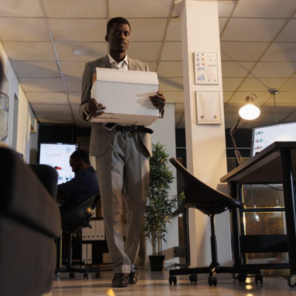 African american entrepreneur holding cardboard box with his belongings after being fired from business startup office. Advisor agent quitting corporate job late at night. Dismissal concept