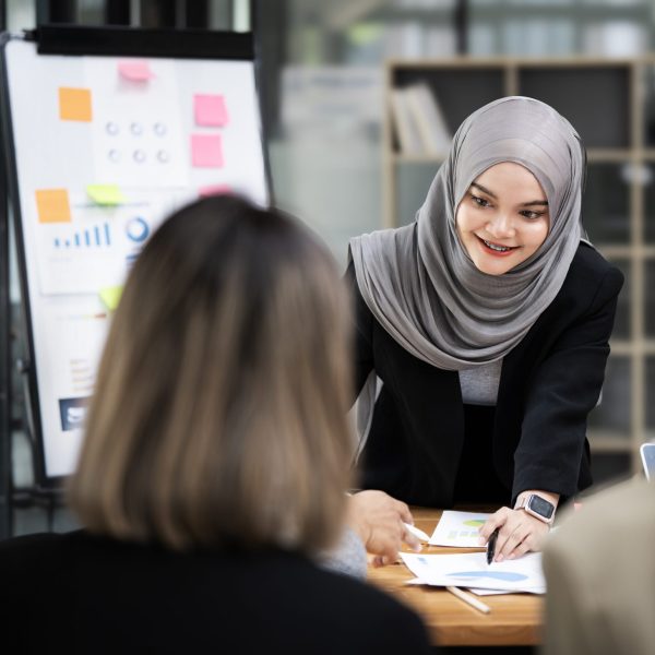 Confident Asian muslim female team leader wearing hijab discussing project statistics with colleagues, standing at table in boardroom