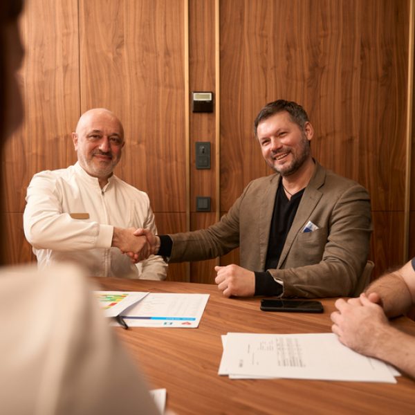 Adult physician sitting at table with colleagues and shaking hands with patient in the office